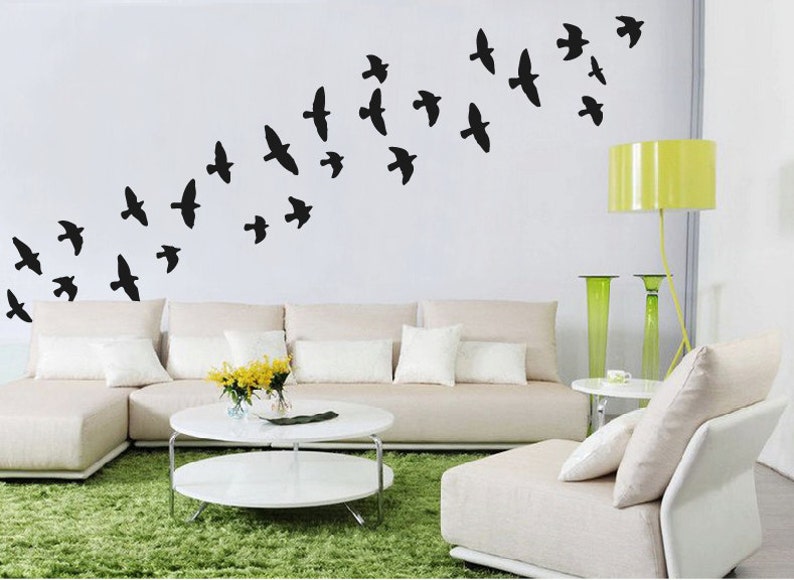 May include: A white couch with white pillows and a white ottoman in front of a white wall with black bird silhouettes. A green rug is on the floor in front of the couch. A yellow lamp is on the right side of the image.