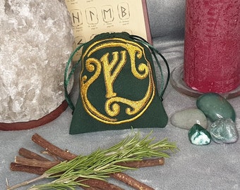 May include: A green pouch with a gold embroidered Yggdrasil symbol, surrounded by rosemary sprigs and licorice sticks.  There are also green and gray stones and a red candle in the background.