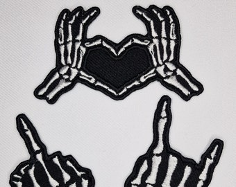 May include: Three iron-on patches featuring skeleton hands in different poses. One patch shows two hands making a heart shape, another shows a hand giving the middle finger, and the last shows a hand giving the rock and roll sign. All patches are black with white skeleton details.