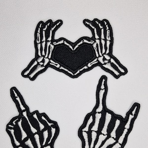 May include: Three iron-on patches featuring skeleton hands in different poses. One patch shows two hands making a heart shape, another shows a hand giving the middle finger, and the last shows a hand giving the rock and roll sign. All patches are black with white skeleton details.