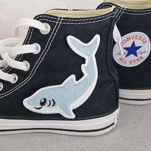 May include: Black high-top Converse trainers with white laces and a blue shark patch on the side. The Converse All Star logo is visible on the side of the shoe.