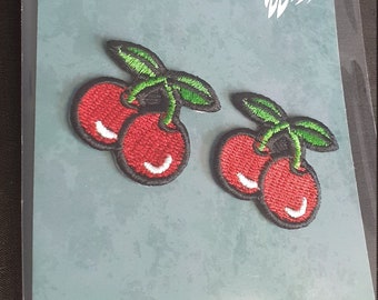 May include: Two embroidered iron-on patches featuring a pair of red cherries with green stems and leaves. The patches are on a blue background with the text "Whyte Raven Crafts" and a raven logo.
