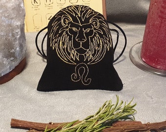 May include: Black velvet pouch with a gold embroidered lion head and the astrological symbol for Leo. The pouch is on a white surface with a salt lamp, a candle, and rosemary sprigs.