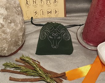 May include: A green velvet pouch with a white embroidered ram's head design. The pouch is surrounded by a variety of objects, including a large salt rock, a red candle, a white bowl, and a bundle of rosemary sprigs.
