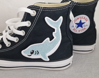 May include: Black high-top Converse sneakers with white laces and a blue shark patch on the side. The Converse All Star logo is visible on the side of the shoe.