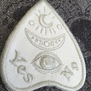 Puede incluir: Parche de fieltro blanco en forma de tri&aacute;ngulo con un dise&ntilde;o bordado en plata. El dise&ntilde;o incluye una luna creciente, un sol, un ojo y una estrella. Las palabras "S&iacute; No Tal vez" est&aacute;n bordadas en la parte inferior del parche.