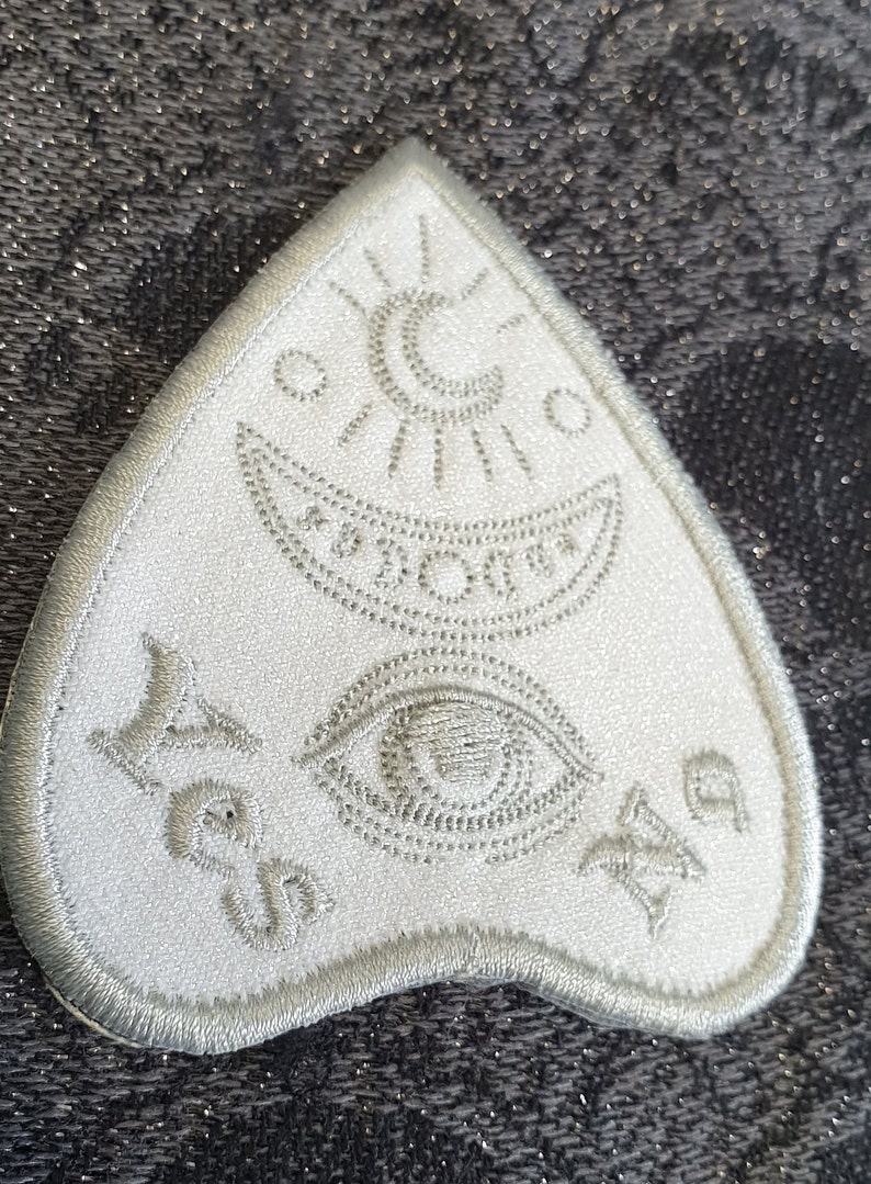 Puede incluir: Parche de fieltro blanco en forma de tri&aacute;ngulo con un dise&ntilde;o bordado en plata. El dise&ntilde;o incluye una luna creciente, un sol, un ojo y una estrella. Las palabras "S&iacute; No Tal vez" est&aacute;n bordadas en la parte inferior del parche.