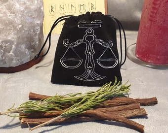 May include: A black velvet pouch with a white embroidered image of a scale, a symbol of balance and justice. The pouch is sitting on a white surface with a sprig of rosemary and licorice sticks.