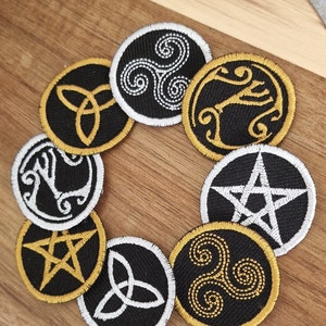 May include: Eight embroidered patches featuring various symbols, including a triquetra, a pentagram, and a triskelion. The patches are black with gold and white embroidery and are arranged in a circle on a wooden surface.