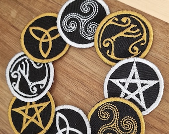 May include: Eight embroidered patches with various symbols, including a triquetra, a pentagram, and a triskelion. The patches are black with gold and white embroidery and are arranged in a circle on a wooden surface.