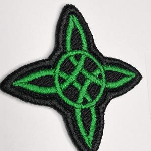 May include: A four-pointed star-shaped embroidered patch featuring a black background with a vibrant green stitched design. The pattern consists of a central circle with intersecting lines that extend into each of the four pointed tips.