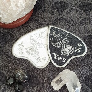 Puede incluir: Un parche de tela blanco y negro con un dise&ntilde;o de luna creciente y sol. El parche est&aacute; dividido en dos, con "S&iacute;" y "No" escritos en cada lado. El parche est&aacute; sobre un fondo de tela negro con cristales blancos.