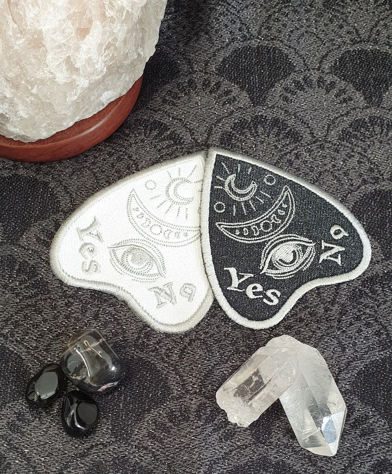 Puede incluir: Un parche de tela blanco y negro con un dise&ntilde;o de luna creciente y sol. El parche est&aacute; dividido en dos, con "S&iacute;" y "No" escritos en cada lado. El parche est&aacute; sobre un fondo de tela negro con cristales blancos.