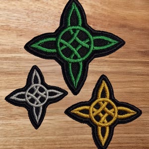 May include: Three embroidered iron-on patches in the shape of a four-pointed star with a circle in the centre. The stars are green, silver, and gold.