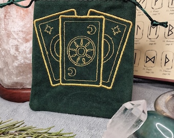 May include: A dark green velvet tarot card bag with a gold embroidered design of three tarot cards. The bag has a drawstring closure and is surrounded by crystals, herbs, and a book with runes.