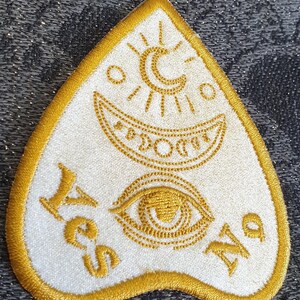 Puede incluir: Un parche de fieltro blanco con un dise&ntilde;o bordado en oro. El dise&ntilde;o presenta una luna creciente, un sol, un ojo y las palabras "S&iacute;" y "No".