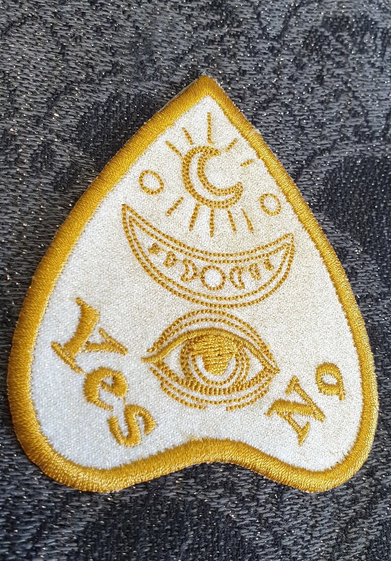 Puede incluir: Un parche de fieltro blanco con un dise&ntilde;o bordado en oro. El dise&ntilde;o presenta una luna creciente, un sol, un ojo y las palabras "S&iacute;" y "No".