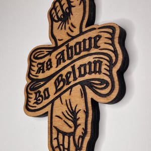 May include: A layered wooden wall art piece featuring a hand pointing upward toward a sunburst and downward toward a crescent moon. A central banner displays the text As Above So Below in black lettering against a natural wood background.