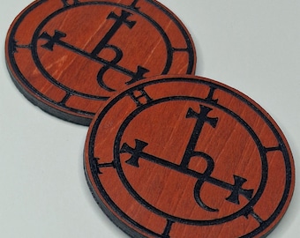 May include: Two circular wooden coasters featuring a reddish-brown finish and a dark, engraved sigil design. Each coaster displays a central cross-like symbol enclosed within a circular border, with black edges and a visible wood grain texture.