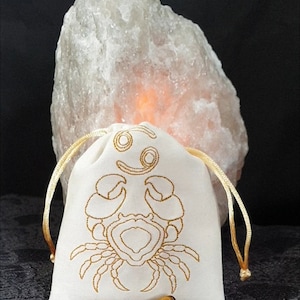 Puede incluir: Una bolsa de cordón blanco con un símbolo del zodíaco Cáncer bordado en oro y un diseño de cangrejo. Dos piedras de ojo de tigre están delante de la bolsa.