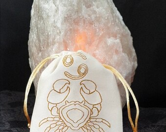 May include: A white drawstring bag with a gold embroidered Cancer zodiac symbol and a crab design. Two tiger's eye stones are in front of the bag.