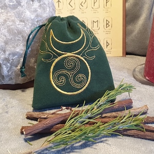 Puede incluir: Una bolsa de terciopelo verde con un diseño de triple espiral y luna creciente bordado en oro. La bolsa está sobre una mesa con palitos de regaliz y una rama de romero.