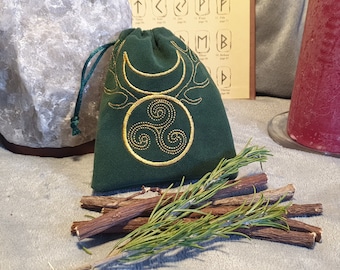 May include: A green velvet pouch with a gold embroidered triple spiral and crescent moon design. The pouch is sitting on a table with sticks of licorice and a sprig of rosemary.