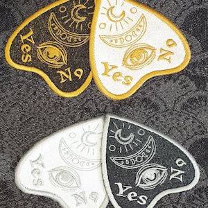 May include: Two black and white planchette patches with gold and silver embroidery. The planchette is shaped like a heart and has the words "Yes" and "No" on it. The planchette is a tool used in Ouija board games.