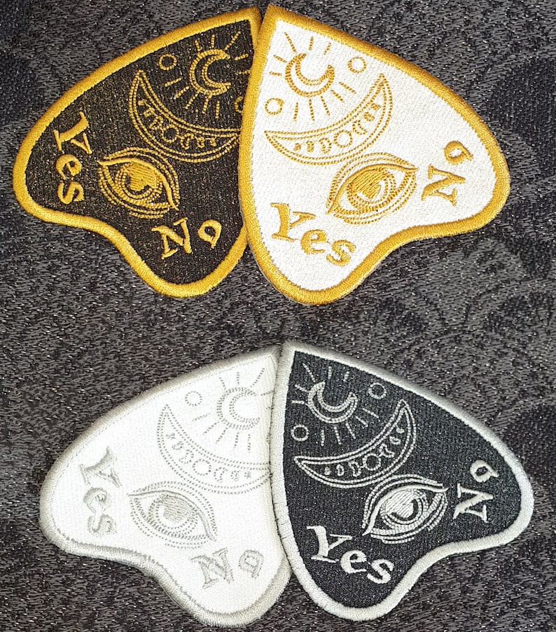 Puede incluir: Dos parches de planchette negros y blancos con bordados dorados y plateados. La planchette tiene forma de coraz&oacute;n y tiene las palabras "S&iacute;" y "No" en ella. La planchette es una herramienta que se utiliza en los juegos de tablero Ouija.