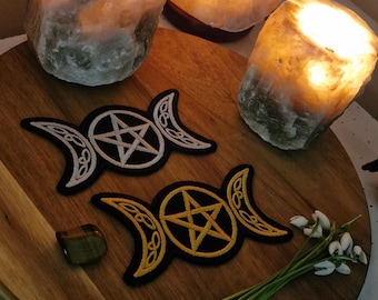 May include: Two embroidered patches featuring a pentagram within a crescent moon design. One patch has a white design on a black background, the other has a yellow design. The patches are on a wooden surface with lit salt lamps.