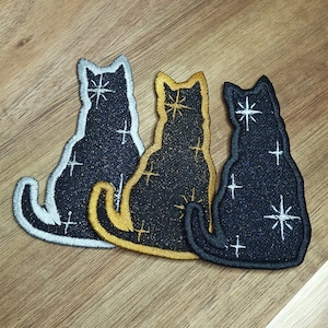 May include: Three embroidered iron-on patches of black cats with a white star design. The cats are in different colours: silver, gold, and black.