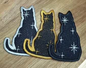 May include: Three embroidered iron-on patches of black cats with a white star design. The cats are in different colors: silver, gold, and black.