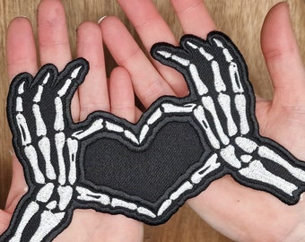 May include: Black fabric patch with two white skeleton hands forming a heart shape. The hands are detailed with bone structure and are stitched onto the fabric.