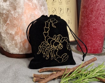 May include: Black velvet pouch with a gold embroidered scorpion design. The pouch has a drawstring closure and is sitting on a gray surface with sticks and rosemary.