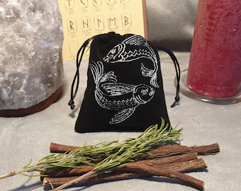 May include: A black velvet pouch with a white embroidered design of two fish. The pouch has a drawstring closure and is surrounded by sprigs of rosemary and licorice sticks.