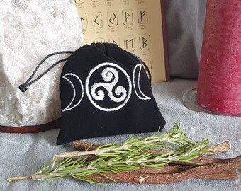 May include: Black velvet pouch with a white embroidered triple moon and triquetra symbol. The pouch is sitting on a gray surface with a sprig of rosemary and sticks.