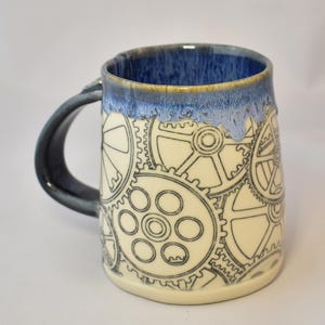 Puede incluir: Una taza de cerámica con cuerpo color crema que presenta un patrón de líneas negras de engranajes mecánicos entrelazados. El borde y el interior están esmaltados en azul profundo con un acabado texturizado y goteante. Un asa azul oscuro.