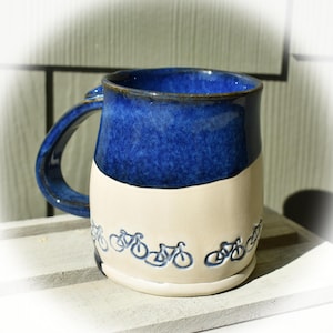Könnte beinhalten: Eine blaue und weiße Keramiktasse mit einem Design von Fahrrädern am Boden. Die Tasse hat einen breiten, abgerundeten Griff.