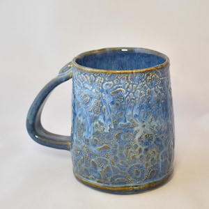 May include: A ceramic mug featuring a textured, lace-like pattern in shades of blue and gold. The mug has a cylindrical body with a curved handle on the side. The glaze shows variations in blue tones with gold accents highlighting the intricate relief.