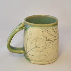 May include: A ceramic mug with a cream-colored body featuring etched maple leaf designs. The handle and the rim are glazed in a variegated green and blue finish, with the green glaze extending down the side of the mug near the handle attachment.