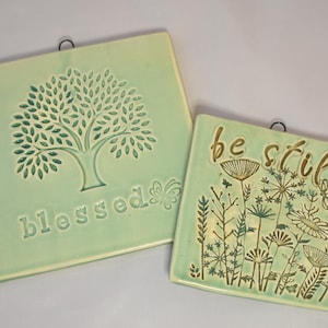 May include: Two square, light green ceramic wall hangings. One features a tree design with the word "blessed" and a butterfly. The other has the words "be still" with a floral design. Both have a metal loop for hanging.