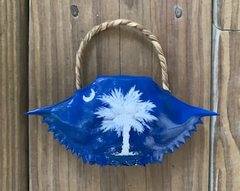 May include: A blue crab shell painted with the South Carolina state flag. The flag features a white palmetto tree and crescent moon on a blue background. The shell has a natural twine loop for hanging.