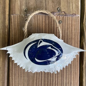 May include: A white crab shell decoration with a navy blue Penn State Nittany Lion logo. The shell is suspended by a natural rope loop against a wooden background. The crab shell has a jagged edge.