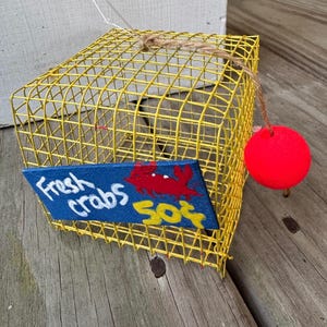 May include: A yellow wire crab trap with a blue sign that reads "Fresh crabs" and a red crab illustration. A red buoy is attached to the trap with twine. The trap is sitting on a wooden surface.