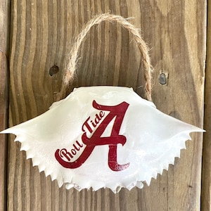 May include: White crab shell ornament with a red "Roll Tide" and the letter "A" painted on it. The ornament has a brown twine hanger.