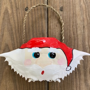 May include: A painted crab shell ornament with a Santa Claus face. The ornament is red, white, and blue and has a rope hanger.