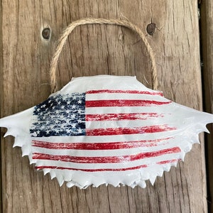 May include: A white crab shell painted with an American flag design. The shell has a twine hanger for hanging.
