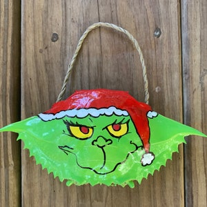 May include: A hand-painted green crab shell ornament featuring a Grinch-like face with a red Santa hat. The ornament has a rope hanger and is set against a wooden background. The Grinch's face has yellow eyes and a mischievous expression.