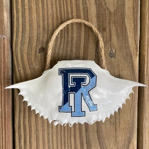 May include: A white crab shell ornament with a blue and navy blue letter "R" painted on it. The shell has a twine loop for hanging.