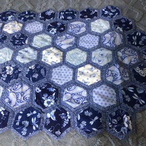 May include: A handmade quilt with a honeycomb design. The quilt is made up of hexagonal patches in shades of blue and white, with floral and paisley patterns. It has a dark blue border around the edges.